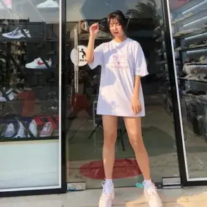Cô gái mang vibe Gấm Kami mặc áo phông trắng dáng rộng đứng trước cửa kính khá tự nhiên