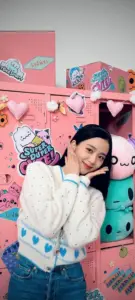 Trong căn phòng hồng, cô gái mặc hoodie trắng đứng cạnh đồ chơi, tạo dáng nhẹ nhàng