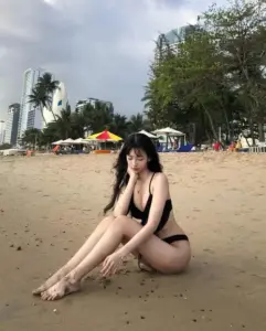Dưới bầu trời nhiều mây, cô mặc bikini đen ngồi trên cát, tóc mái che nhẹ