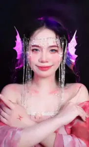 Màn đèn tím làm nổi cô trang điểm lấp lánh, đan tay trước ngực cười nhẹ