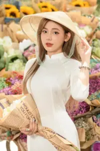 Nàng mặc áo dài trắng đội nón lá, nhìn thẳng ống kính giữa gian hàng hoa rực màu