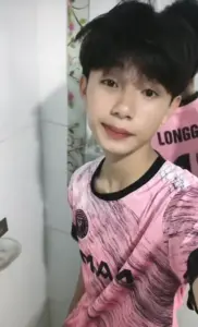 Cậu bạn áo hồng đứng trong phòng, tóc đen phồng nhẹ và nét mặt dễ thương đúng tuổi